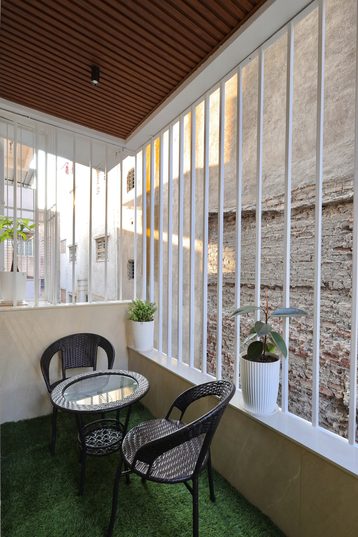 A narrow balcony behind white vertical bars, with a slatted wooden ceiling, artificial grass, a glass-topped table and two woven chairs.