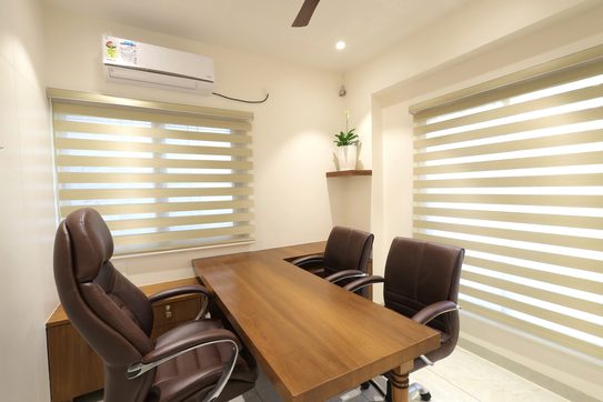 An L-shaped desk and three leather chairs in a corner cabin, zebra blinds on both windows and a small shelf with a plant between them.