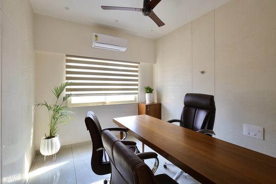 A cabin with a long wooden table and brown leather chairs, a window with zebra blinds at the far end and a palm in a white fluted pot catching the sun.