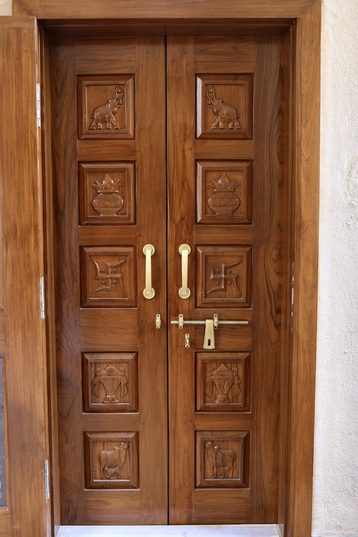A double door in dark wood, each leaf carved in five panels with elephants, kalash and cows, with brass pull handles and a brass latch.