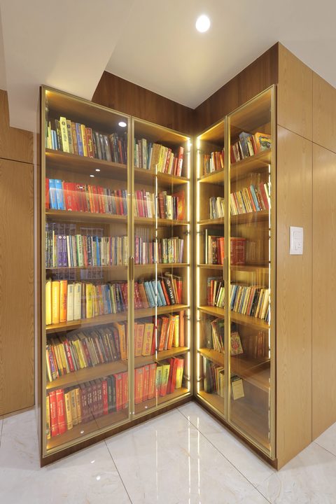 A glass-fronted bookcase that turns a corner, lit along its uprights and full of books, set into wood-grain panelling.