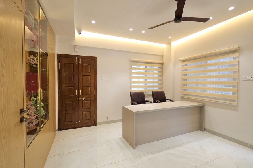 The reception: a pale desk with two leather chairs, a dark panelled double door, zebra blinds on two windows and a cove of warm light around the ceiling, with a lit glass display case set into a wood-grain wall on the left.