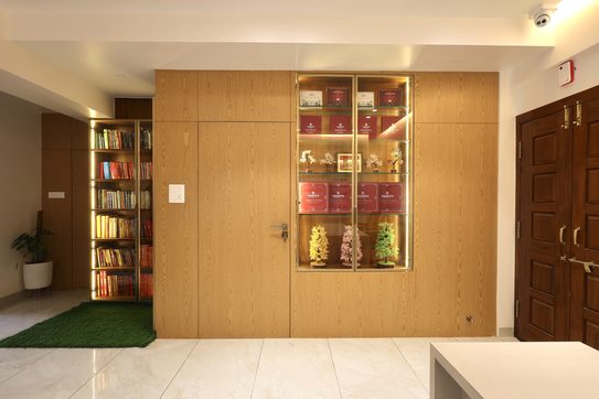 A full-height wood-grain wall with a lit glass display case set into it and a flush door that shows only its handle; a lit bookcase in the passage to the left, the double door to the right.