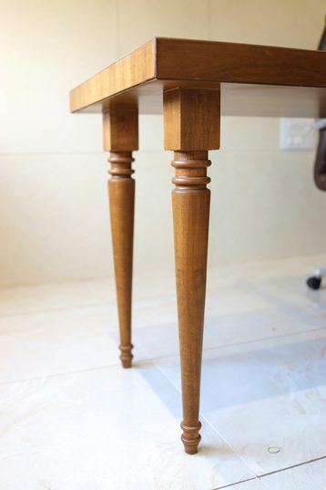 The corner of a long table, a thick wooden top on turned, tapering legs.