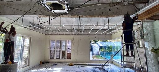 Two workers on ladders fixing a false ceiling frame in a bare room with unglazed windows.